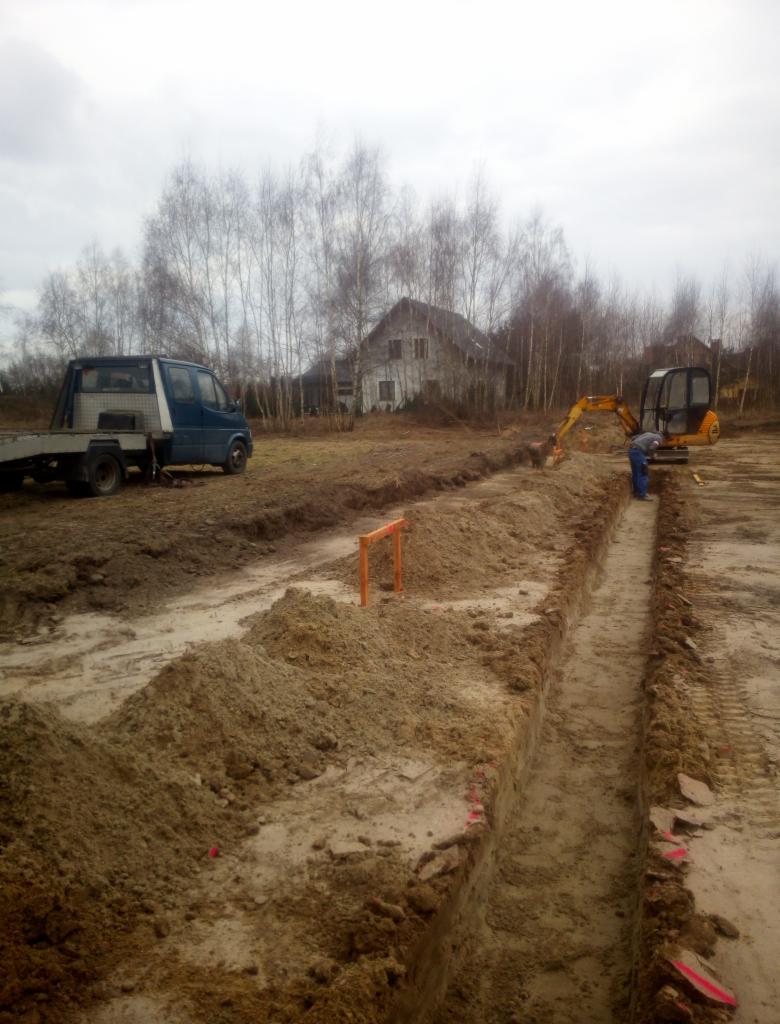 Mała koparka wykopuje wąski rów w ziemi, obok zaparkowany samochód dostawczy, w tle dom i drzewa bez liści.