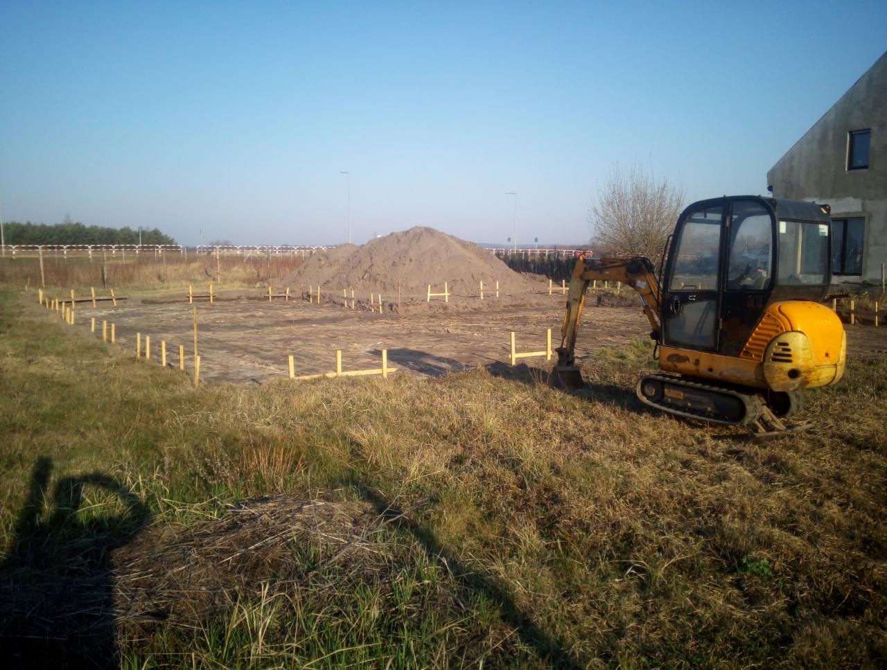 Żółta mini koparka stoi na działce z wyznaczonym obrysem fundamentów pod budynek, w tle pryzma ziemi i budynek w trakcie budowy.