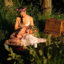 Boskie Foto - Dwie kobiety w wiankach z kwiatów siedzą na kocu w trawie podczas pikniku, obok kosz wiklinowy i truskawki.