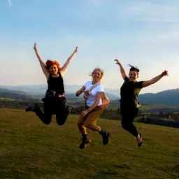 Trzy kobiety w różnym wieku skaczą radośnie na wzgórzu z widokiem na malowniczy krajobraz górski w ciepłym świetle zachodzącego słońca.