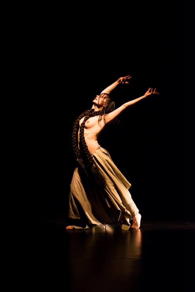 Kobieta w ruchu tanecznym, oświetlona punktowo na czarnym tle, z bardzo długim, grubym warkoczem opadającym na tors i zwiewną spódnicą.