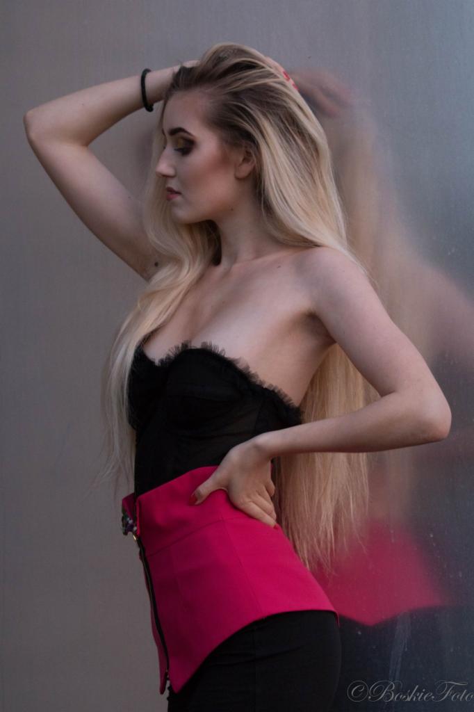 Portret kobiety z blond włosami, w czarnym gorsecie i fuksjowej spódnicy, opierającej się o szarą ścianę z odbiciem, wizaż z ciemnym cieniem do powiek, lekko rozchylone usta.