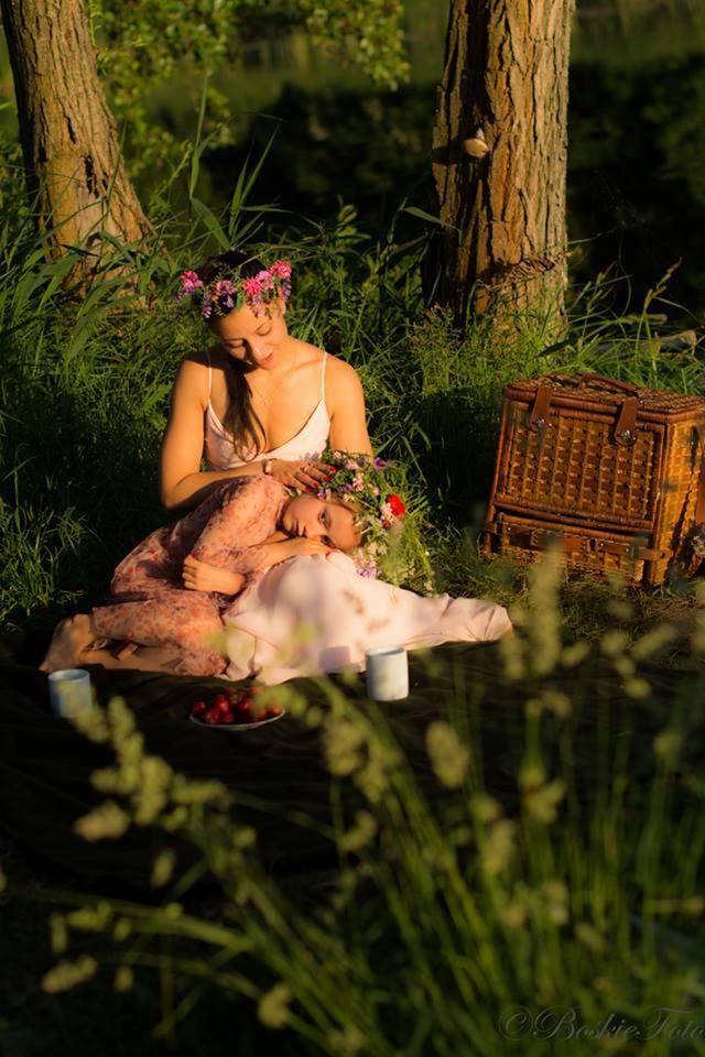 Dwie kobiety w wiankach z kwiatów siedzą na kocu w trawie podczas pikniku, obok kosz wiklinowy i truskawki.