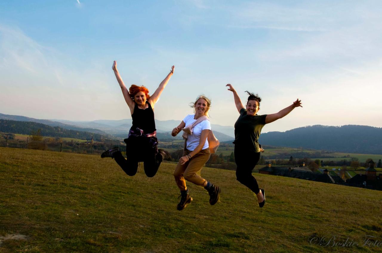 Trzy kobiety w różnym wieku skaczą radośnie na wzgórzu z widokiem na malowniczy krajobraz górski w ciepłym świetle zachodzącego słońca.