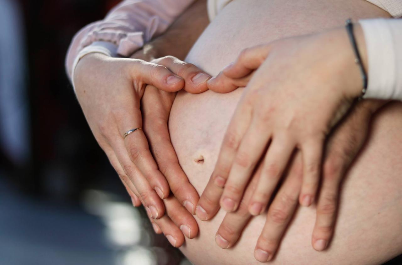 Dłonie kobiety z pierścionkiem i mężczyzny układają się w kształt serca na ciążowym brzuszku, delikatne oświetlenie.