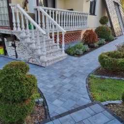 Kaskadowe schody wejściowe z białymi balustradami, prowadzące do domu z szarą kostką brukową, otoczone równo przyciętymi krzewami i rabatami kwiatowymi, z zapasem kostki pod schodami.