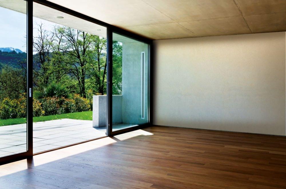 Nowoczesne, przesuwne okno tarasowe z czarnymi ramami, otwarte na taras z widokiem na zieleń, drewniana podłoga wewnątrz pomieszczenia, betonowy sufit.