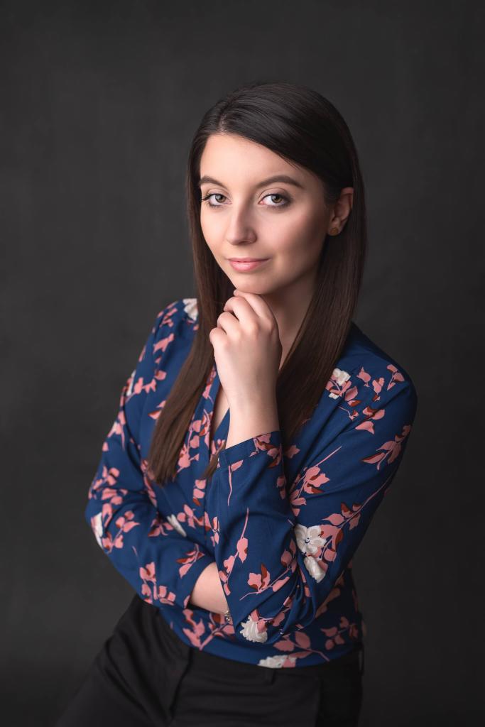 Portret kobiety z ciemnymi włosami w niebieskiej bluzce w kwiaty, opierającej brodę na dłoni, na ciemnoszarym tle.