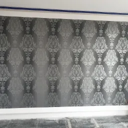 Ściana pokryta tapetą w odcieniach szarości z motywem roślinnym w stylu retro, widoczne niedoskonałości wykończenia przy podłodze.