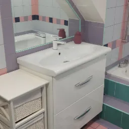 Umywalka z szafką w łazience z ukośnym sufitem, widoczne odbicie w lustrze, kolorowe płytki podłogowe.