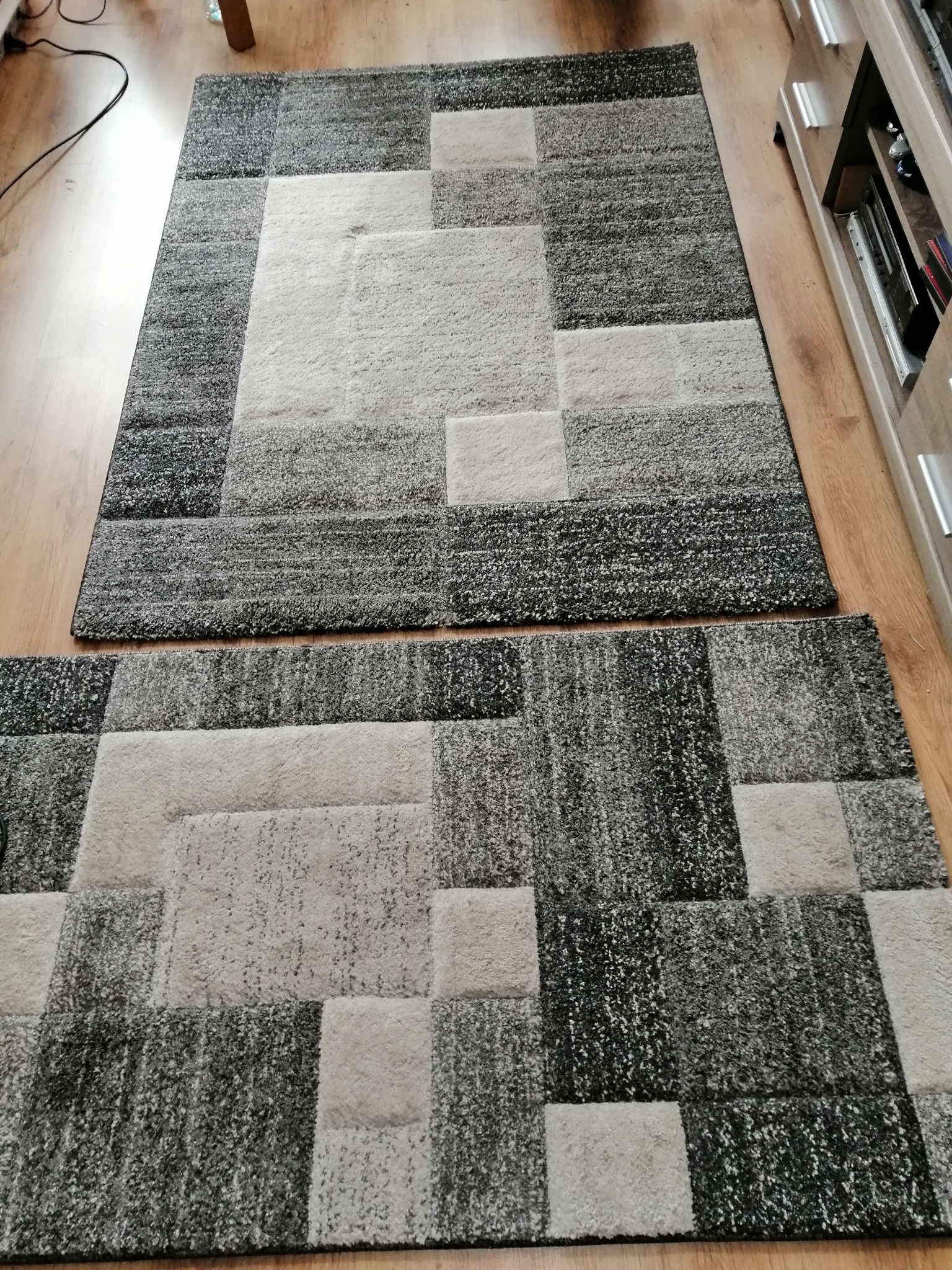 Dwa prostokątne dywany z geometrycznym wzorem w odcieniach szarości i beżu, ułożone na drewnianej podłodze, widoczne z góry.