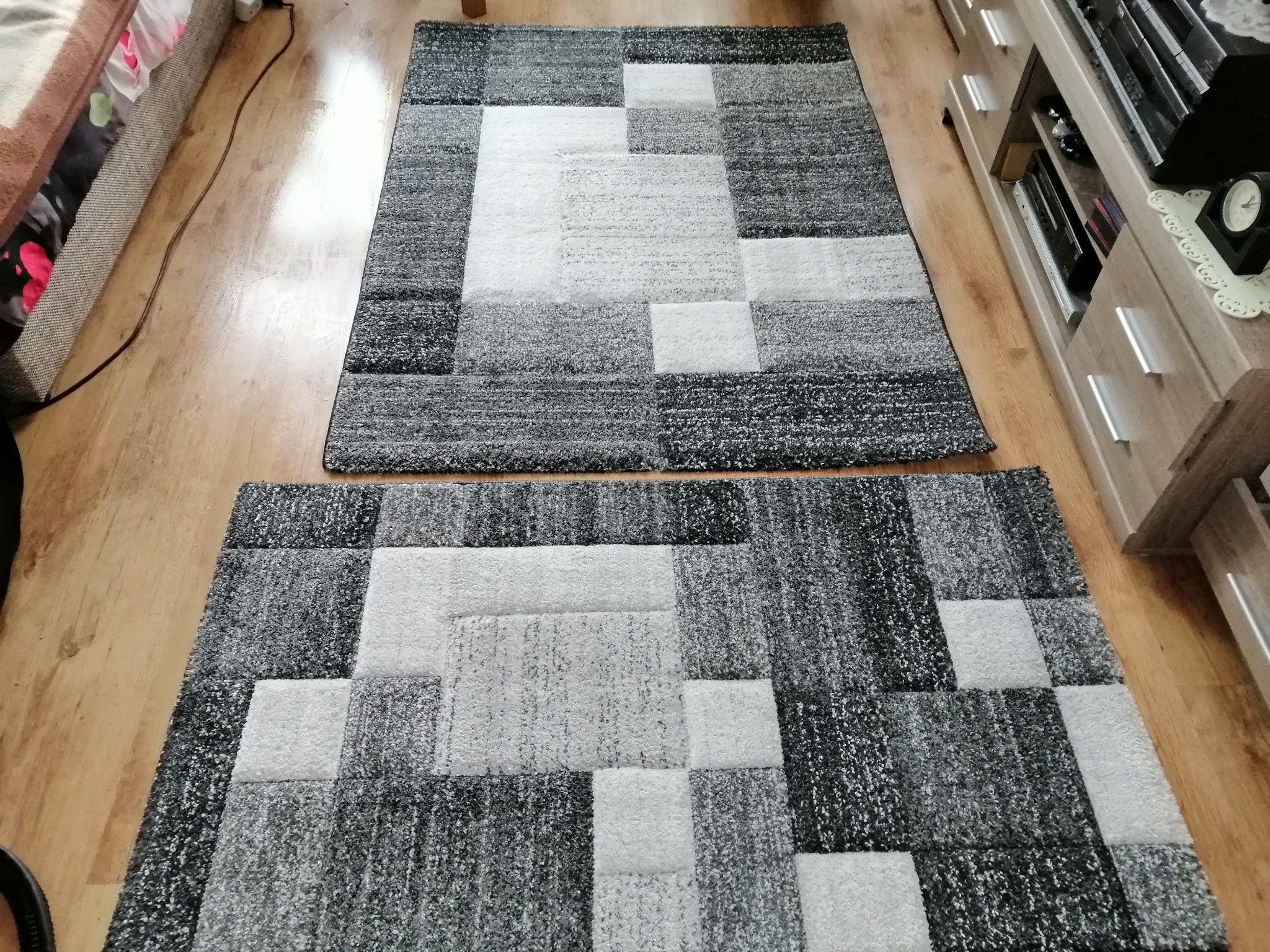 Dwa identyczne dywany w geometryczne wzory (szaro-białe kwadraty) leżące na drewnianej podłodze, widok z góry.