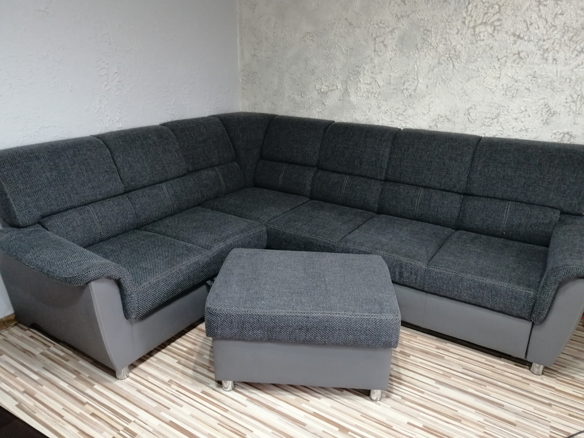 Szara, narożna kanapa z pufą na tle ściany z dekoracyjnym tynkiem, ustawiona na podłodze z paneli imitujących drewno.