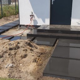 Usługi budowlano remontowe RYSZARD HĘCIAK - Montaż ścieżki z ciemnych, kwadratowych płyt kamiennych prowadzącej do wejścia domu z widocznym stosem piasku i elementami instalacji wodnej na elewacji.