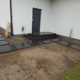 Usługi budowlano remontowe RYSZARD HĘCIAK - Układanie czarnych, kwadratowych płyt kamiennych jako ścieżki prowadzącej do drzwi wejściowych domu, widoczne etapy prac ziemnych i przygotowania podłoża.