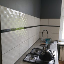 Usługi budowlano remontowe RYSZARD HĘCIAK - Ściana w kuchni z ułożonymi białymi płytkami z delikatną fakturą, oddzielonymi czarnym pasem, nad blatem z kuchenką gazową, na której stoi czajnik, obok widoczne narzędzia i krzyżyki do płytek.