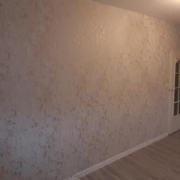 Usługi budowlano remontowe RYSZARD HĘCIAK - Wykończone wnętrze pokoju z dekoracyjną ścianą w odcieniach beżu i brązu, drewnopodobna podłoga, białe drzwi i fragment okna z firanką z kwiatowym motywem.