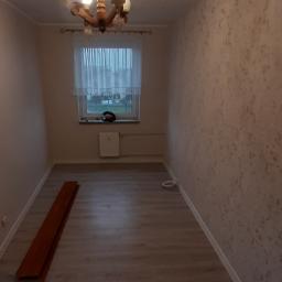 Usługi budowlano remontowe RYSZARD HĘCIAK - Pusty pokój z oknem, drewnianym żyrandolem i panelami podłogowymi w trakcie montażu. Na ścianie widoczna strukturalna tapeta w beżowych odcieniach.