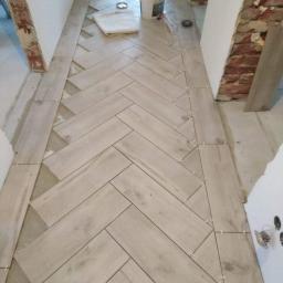 Usługi budowlano remontowe RYSZARD HĘCIAK - Korytarz w trakcie układania podłogi z płytek imitujących drewno w jodełkę, widoczne krzyżyki dystansowe, wiadro z klejem i odsłonięta cegła na ścianie.