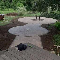 Okrągły plac z kostki brukowej z miejscem na ognisko, połączony ścieżką z kostki, widok z drewnianego tarasu, narzędzia i ubranie leżą na tarasie.