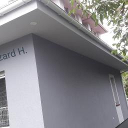 Usługi budowlano remontowe RYSZARD HĘCIAK - Szara elewacja budynku z widocznym napisem firmy 'UBR Ryszard H.', białe okno z firankami i białe rynny, w tle drzewa i sterta drewna.