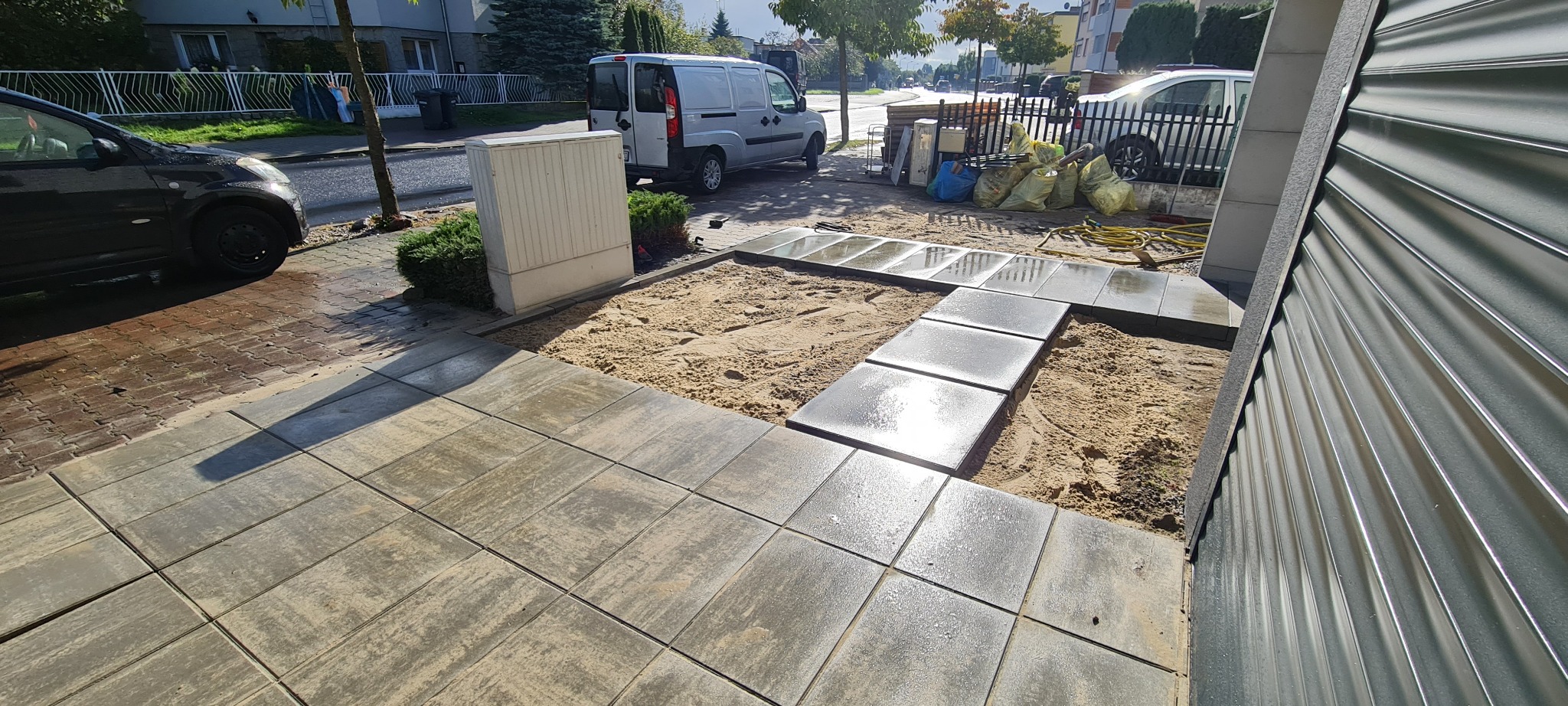 Ułożona kostka brukowa na podjeździe z widocznym fragmentem ścieżki z płyt betonowych, piasek wyrównujący podłoże, zaparkowany bus i samochód osobowy w tle.