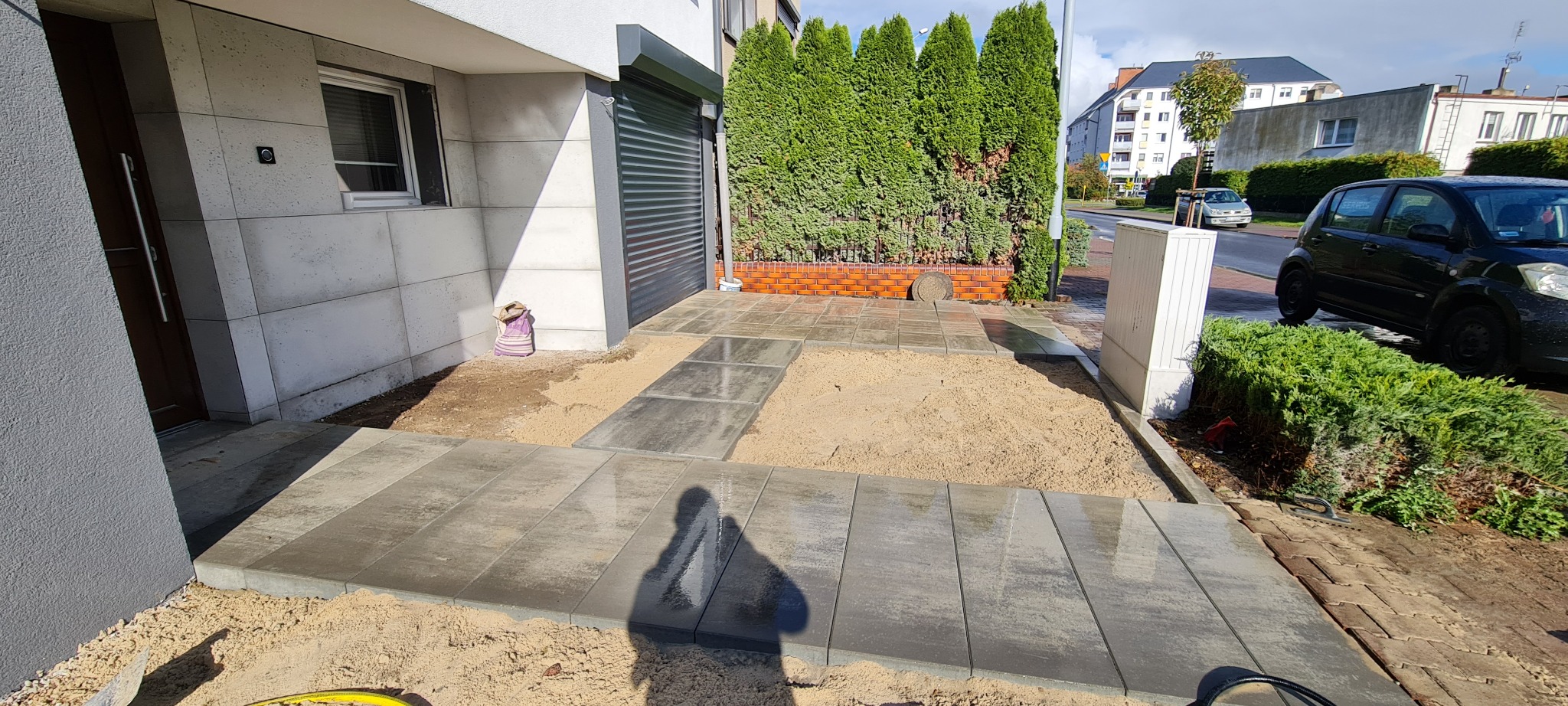 Fragment podjazdu z ułożoną częścią szarej kostki brukowej, widoczny piasek pod kostką, cień osoby fotografującej, w tle budynek z garażem i zielone tuje.