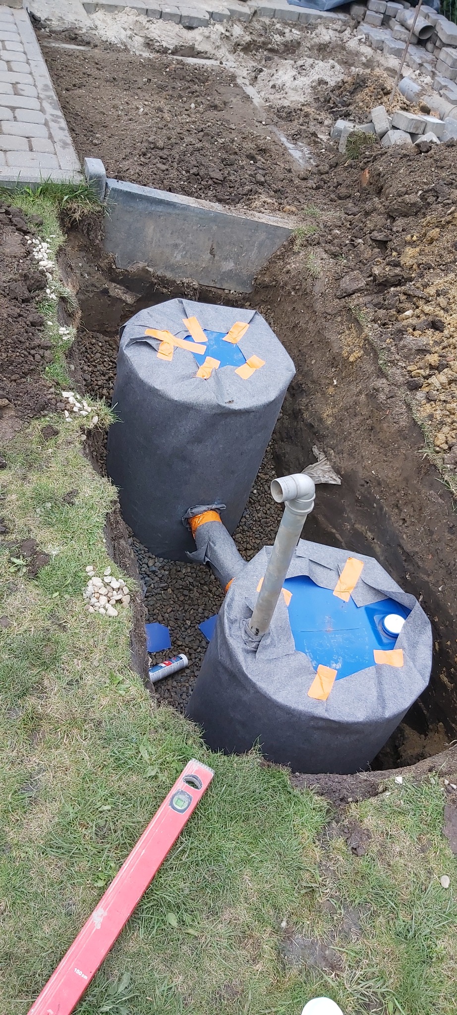 Instalacja dwóch zbiorników retencyjnych w wykopie, zabezpieczonych szarą geowłókniną i pomarańczową taśmą, z widocznymi rurami łączącymi, poziomica na trawie i częściowo ułożona kostka brukowa w tle.