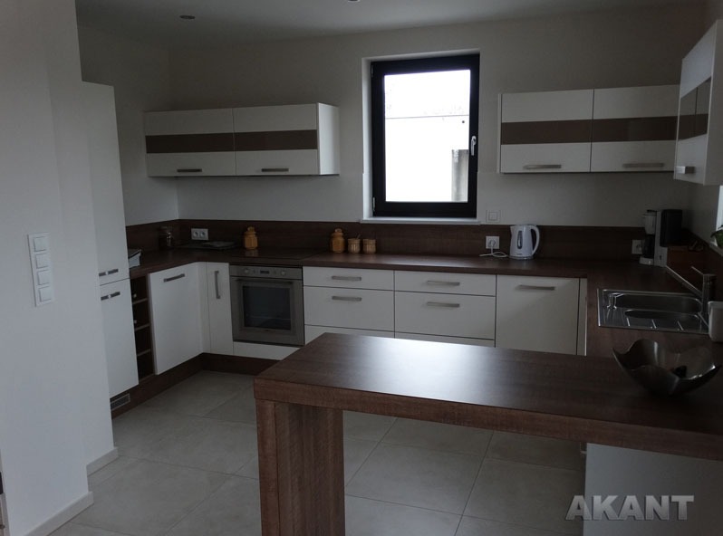 Nowoczesna kuchnia z białymi szafkami z brązowymi akcentami, drewnianym blatem i wyspą kuchenną z ciemnego drewna, oświetlona naturalnym światłem z okna.