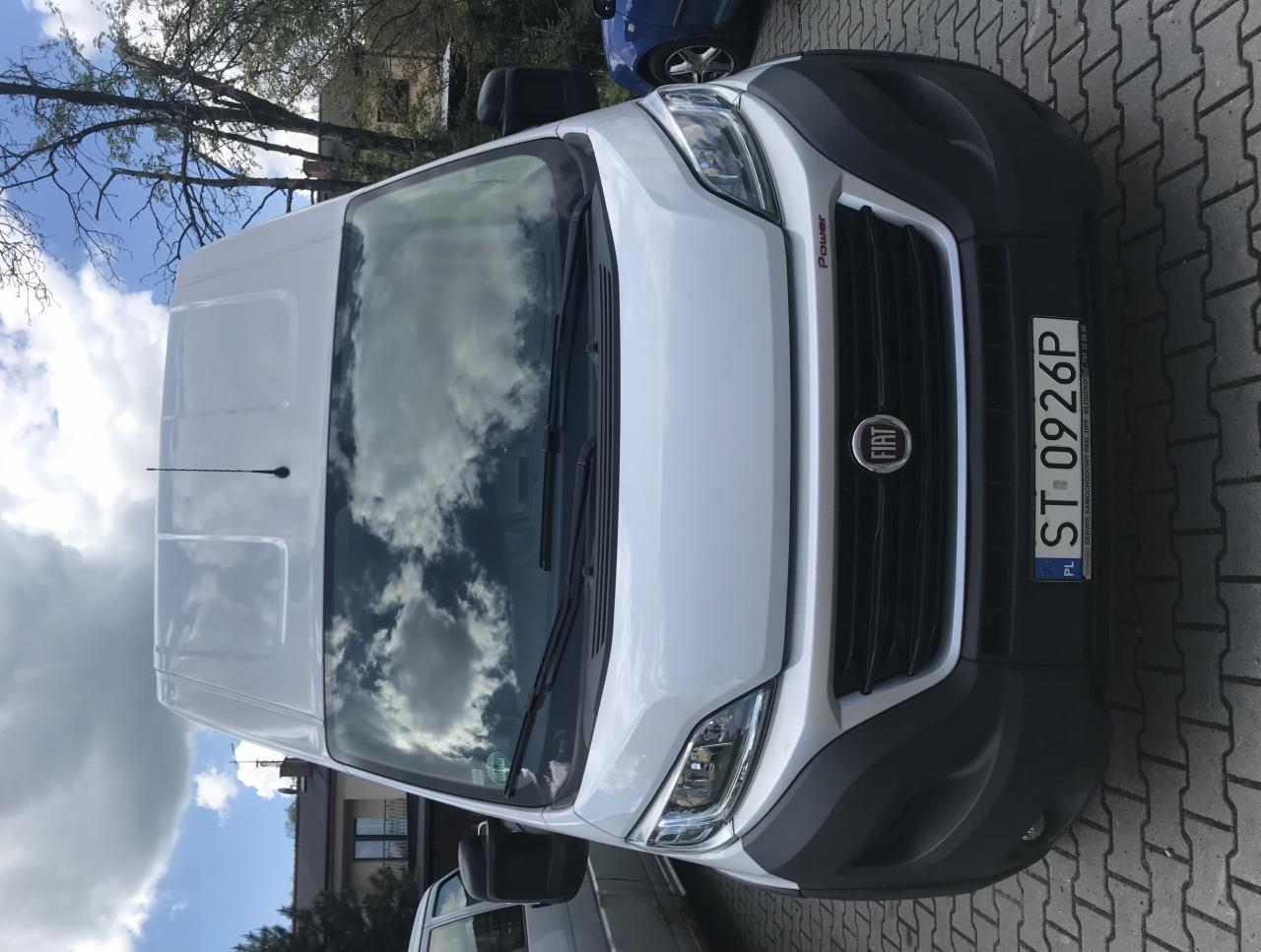 Biały samochód dostawczy Fiat Ducato z polską tablicą rejestracyjną ST 0926P, zaparkowany na brukowanej powierzchni, z widocznym odbiciem chmur na przedniej szybie.