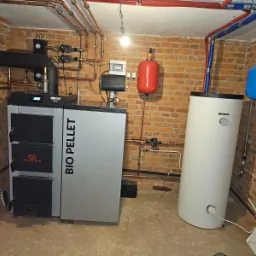 Kotłownia z kotłem na pellet Bio Pellet, zasobnikiem ciepłej wody użytkowej i instalacją grzewczą z rur miedzianych na tle ceglanej ściany.