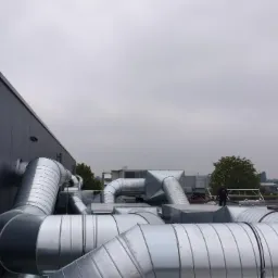 Srebrne rury wentylacyjne na dachu budynku przemysłowego, widoczne elementy systemu rekuperacji i pracownik w oddali, pochmurne niebo.