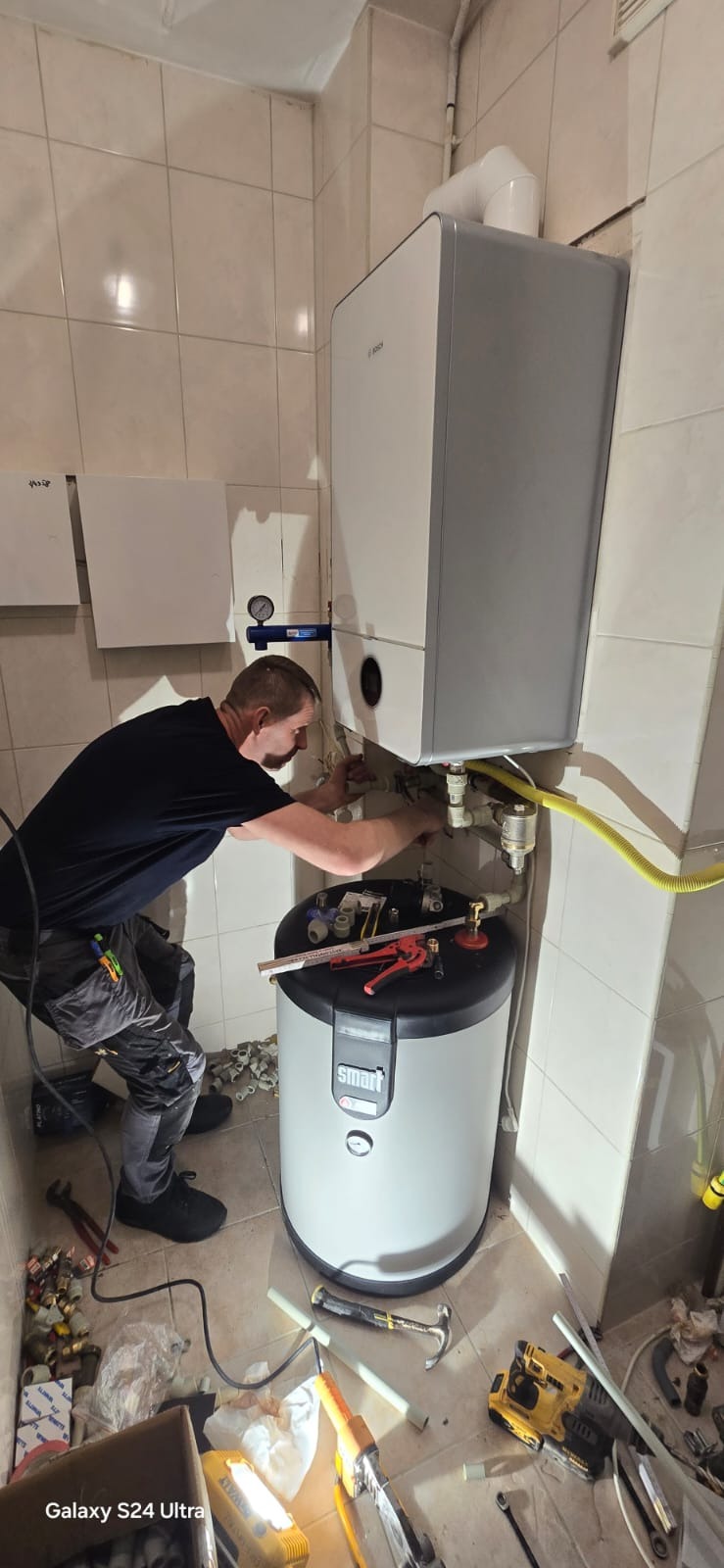 Hydraulik w trakcie montażu dwufunkcyjnego pieca gazowego i zasobnika wody w łazience wyłożonej kafelkami, widoczne narzędzia i elementy instalacji.