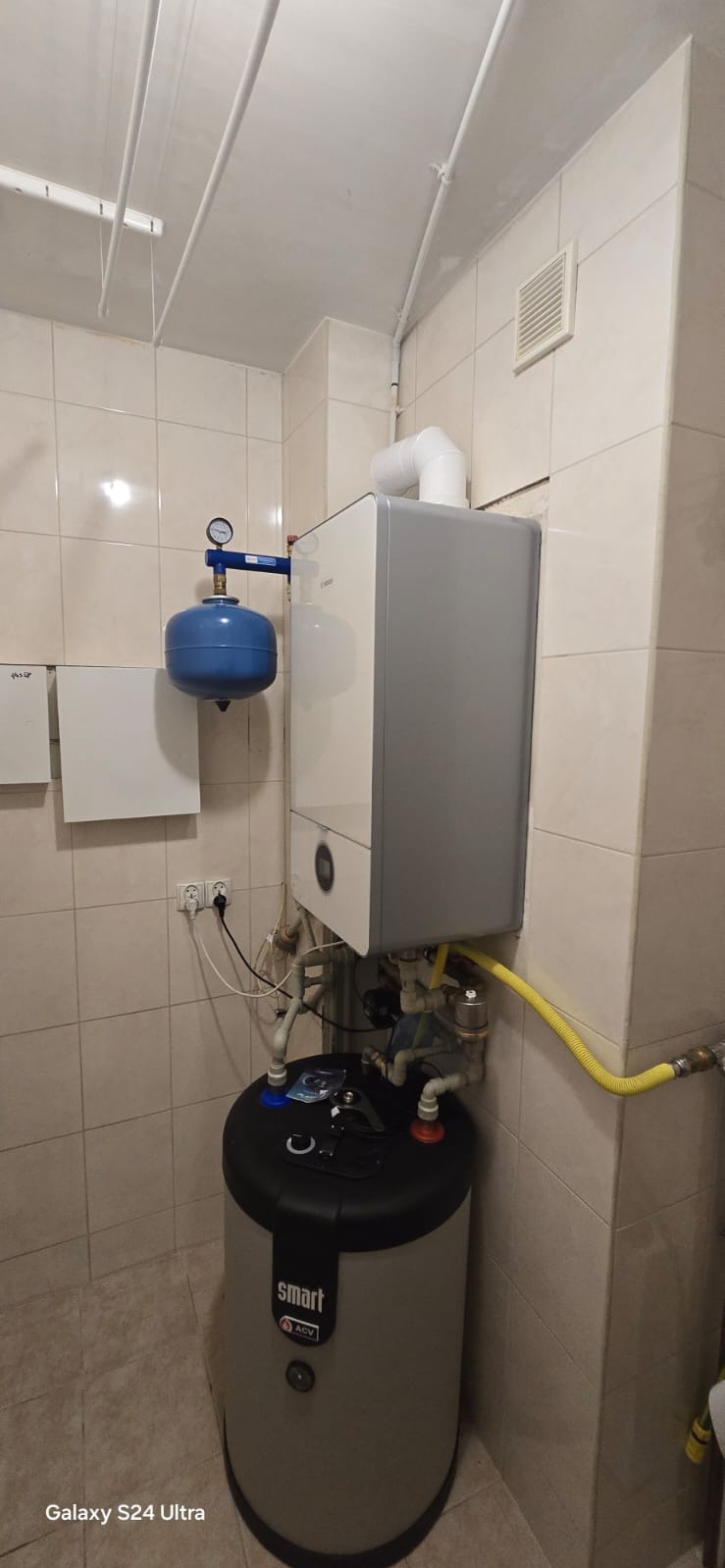 Nowoczesny piec gazowy kondensacyjny zamontowany w pomieszczeniu z beżowymi płytkami, widoczny zbiornik buforowy ACV Smart oraz naczynie przeponowe z manometrem.