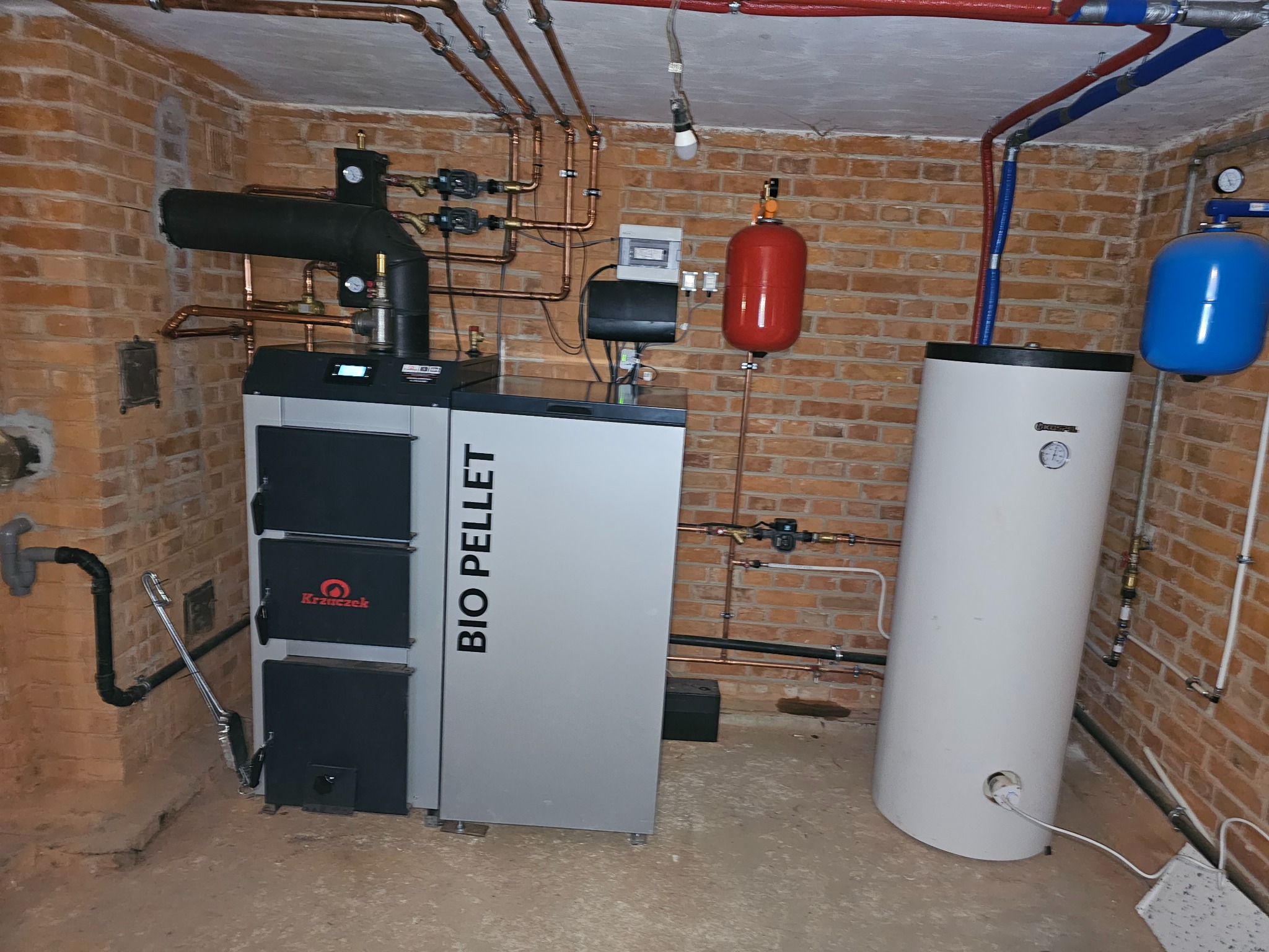 Kotłownia z piecem na pellet marki Krzaczek, zbiornikiem buforowym i instalacją grzewczą z rur miedzianych w pomieszczeniu z ceglanymi ścianami.