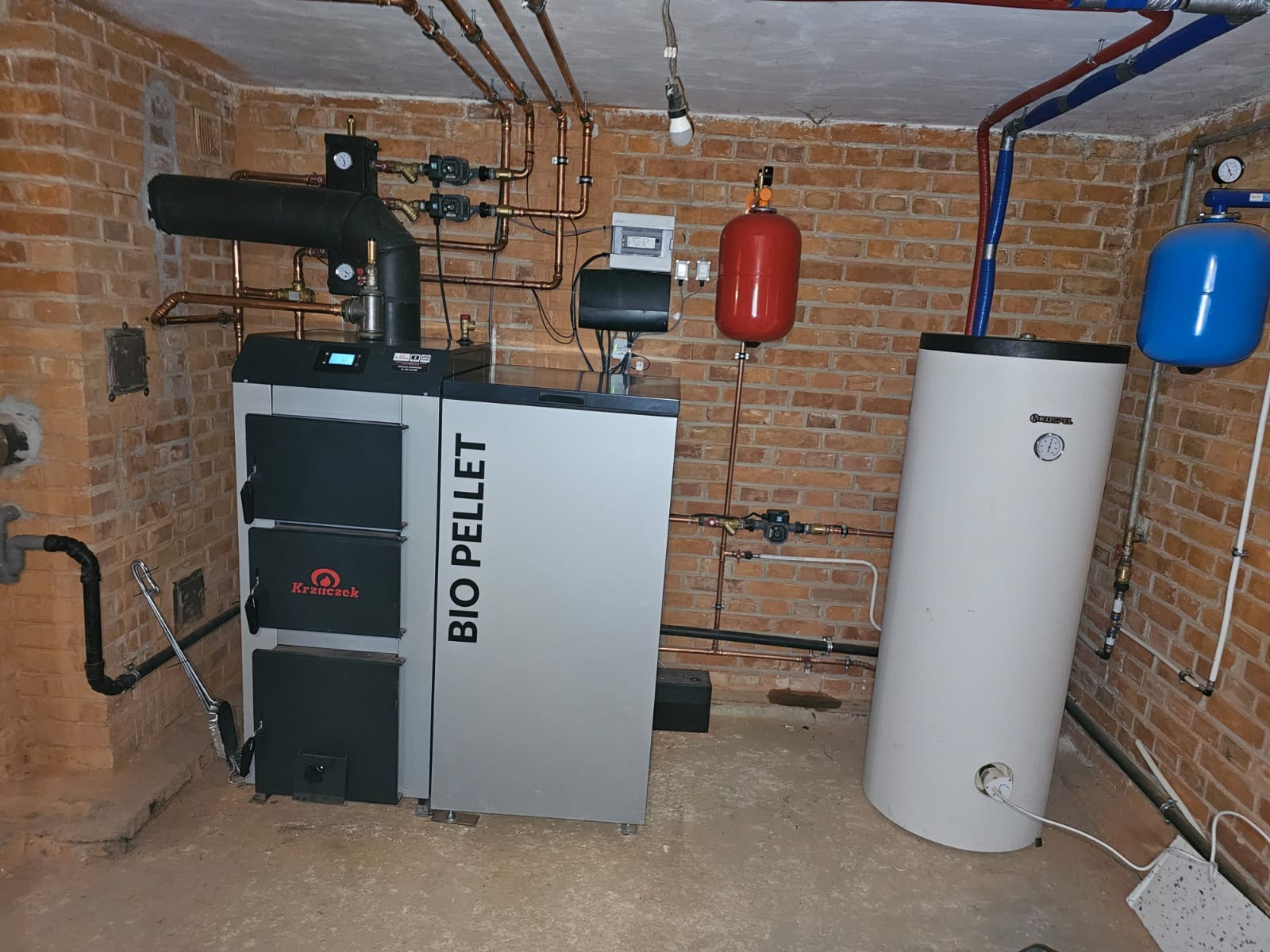 Kotłownia z kotłem na pellet marki Krzaczek, zbiornikiem akumulacyjnym Kospel i instalacją z miedzianych rur na tle ściany z czerwonej cegły.