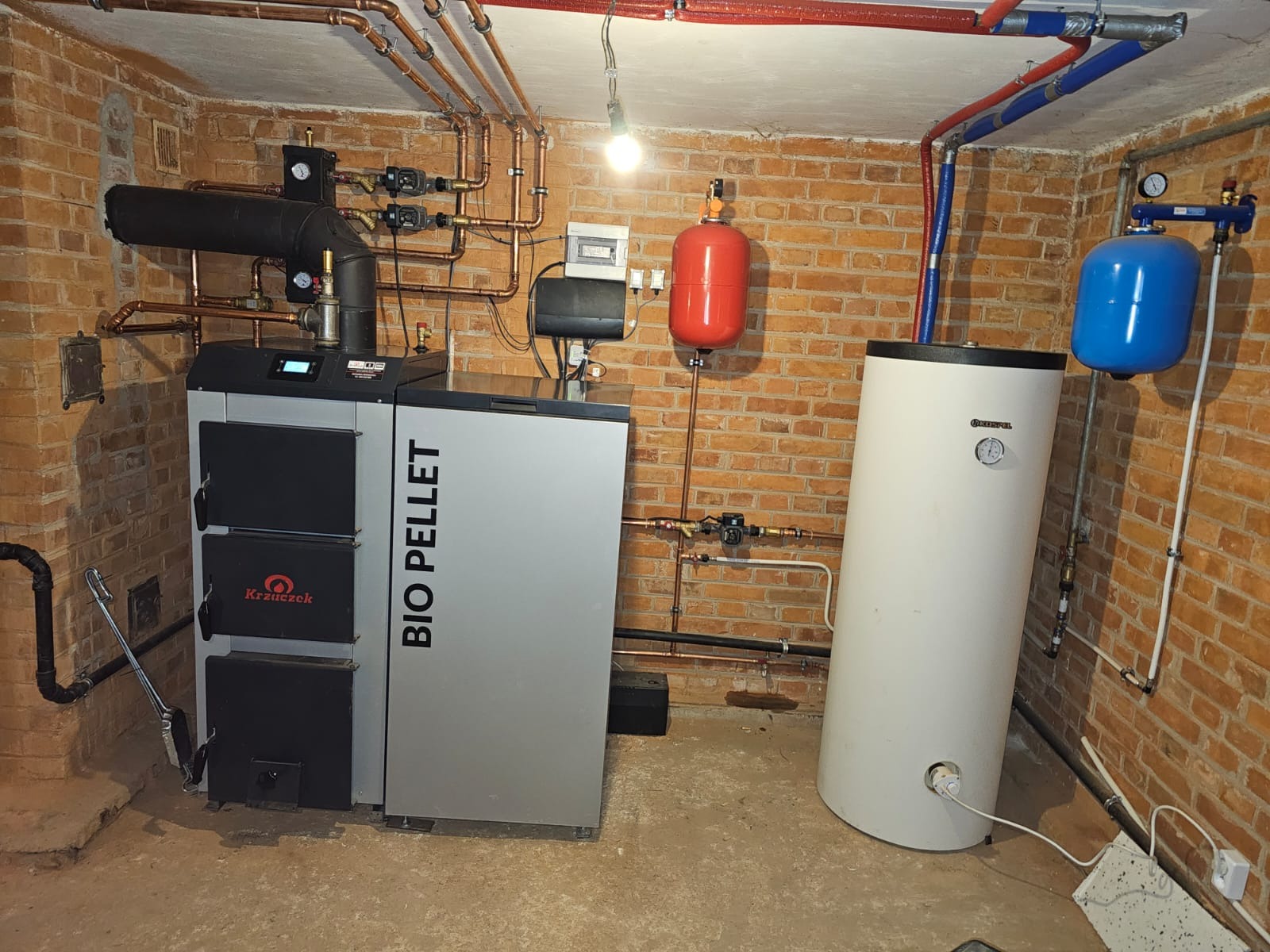 Kotłownia z kotłem na pellet Bio Pellet, zasobnikiem ciepłej wody użytkowej i instalacją grzewczą z rur miedzianych na tle ceglanej ściany.