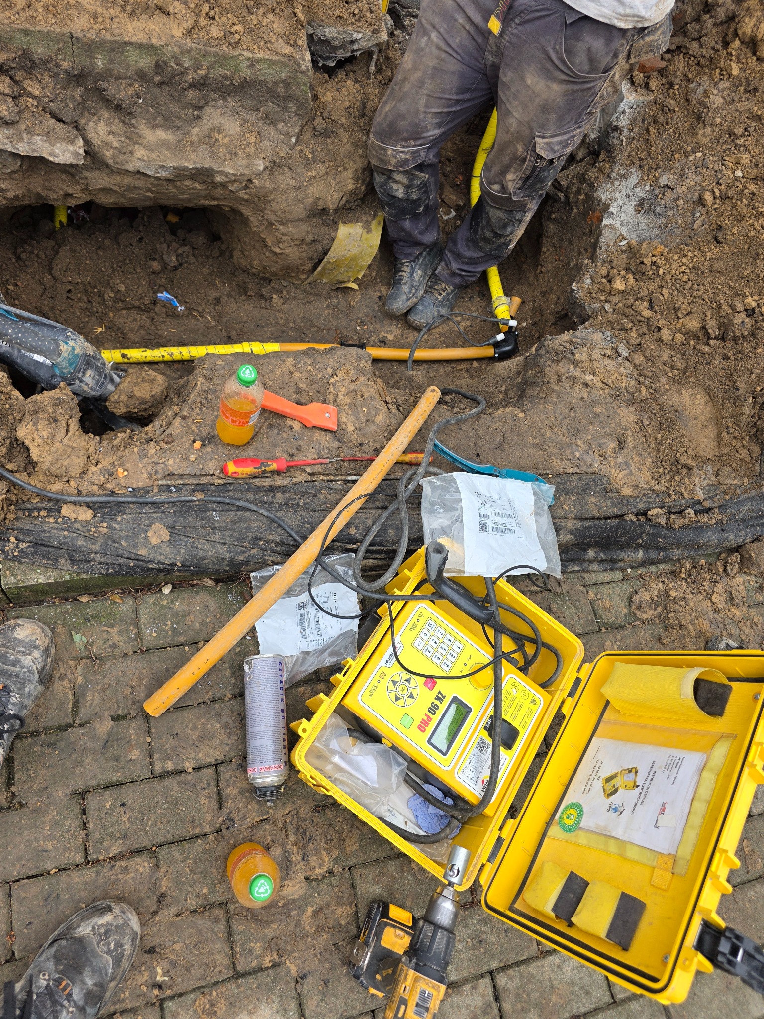 Wykop z żółtymi rurami gazowymi, narzędzia i detektor nieszczelności ZK 90 PRO. Prace ziemne i instalacja gazowa w trakcie realizacji. Widoczne elementy przyłącza.