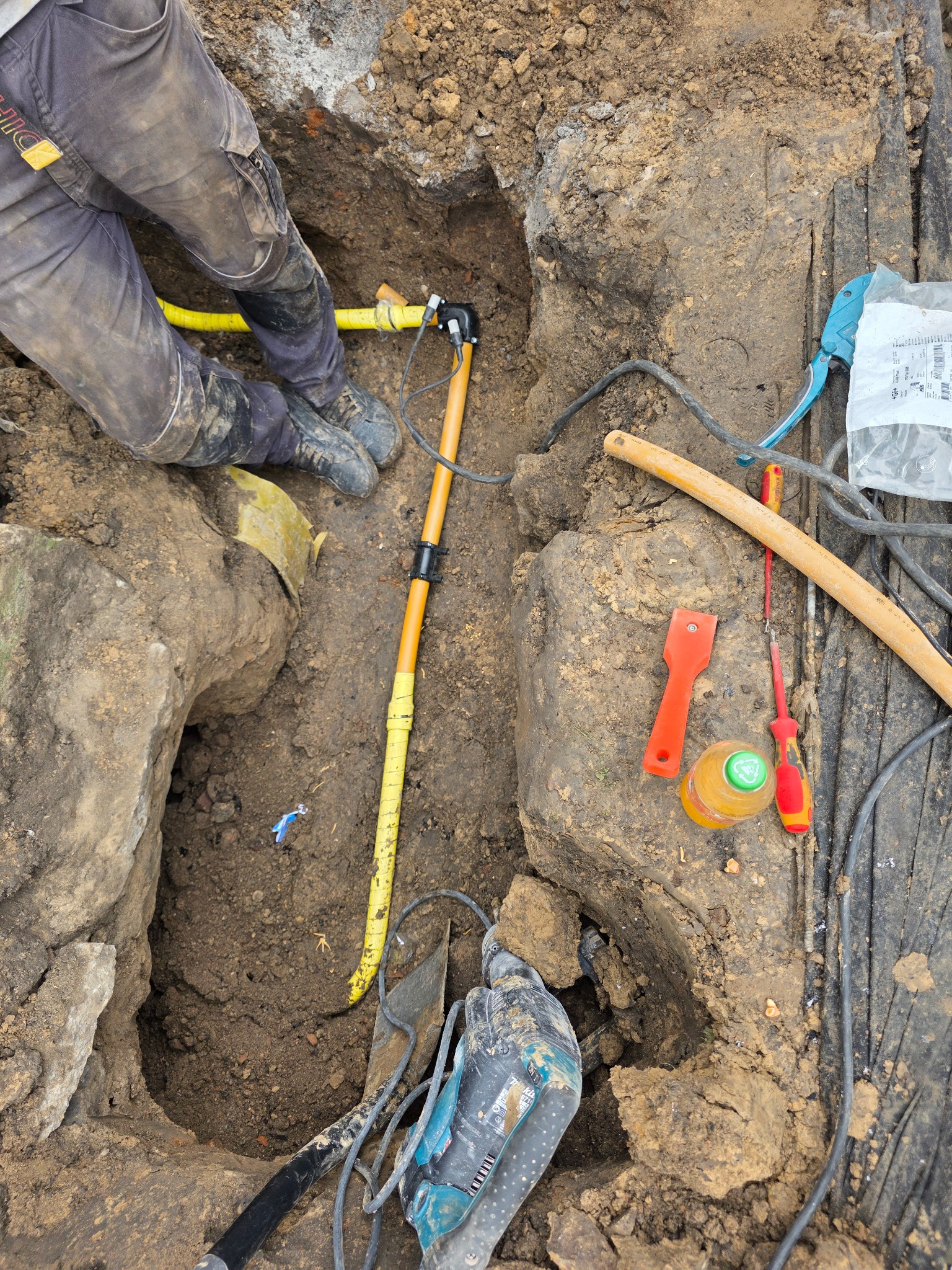 Żółta rura gazowa w wykopie ziemnym, widoczne narzędzia i fragment osoby w kombinezonie roboczym. Wykonywanie przyłącza gazowego.