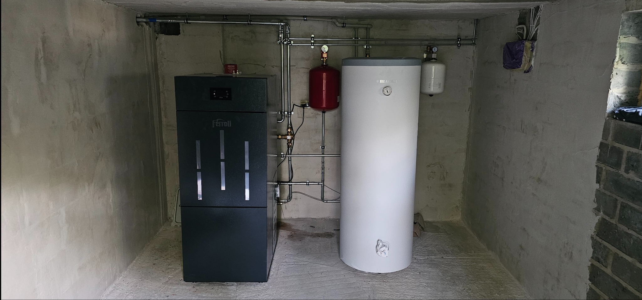 Zestaw grzewczy: piec Ferroli, biały zasobnik Biawar i czerwony zbiornik wyrównawczy, połączone rurami w surowym pomieszczeniu z betonowymi ścianami.