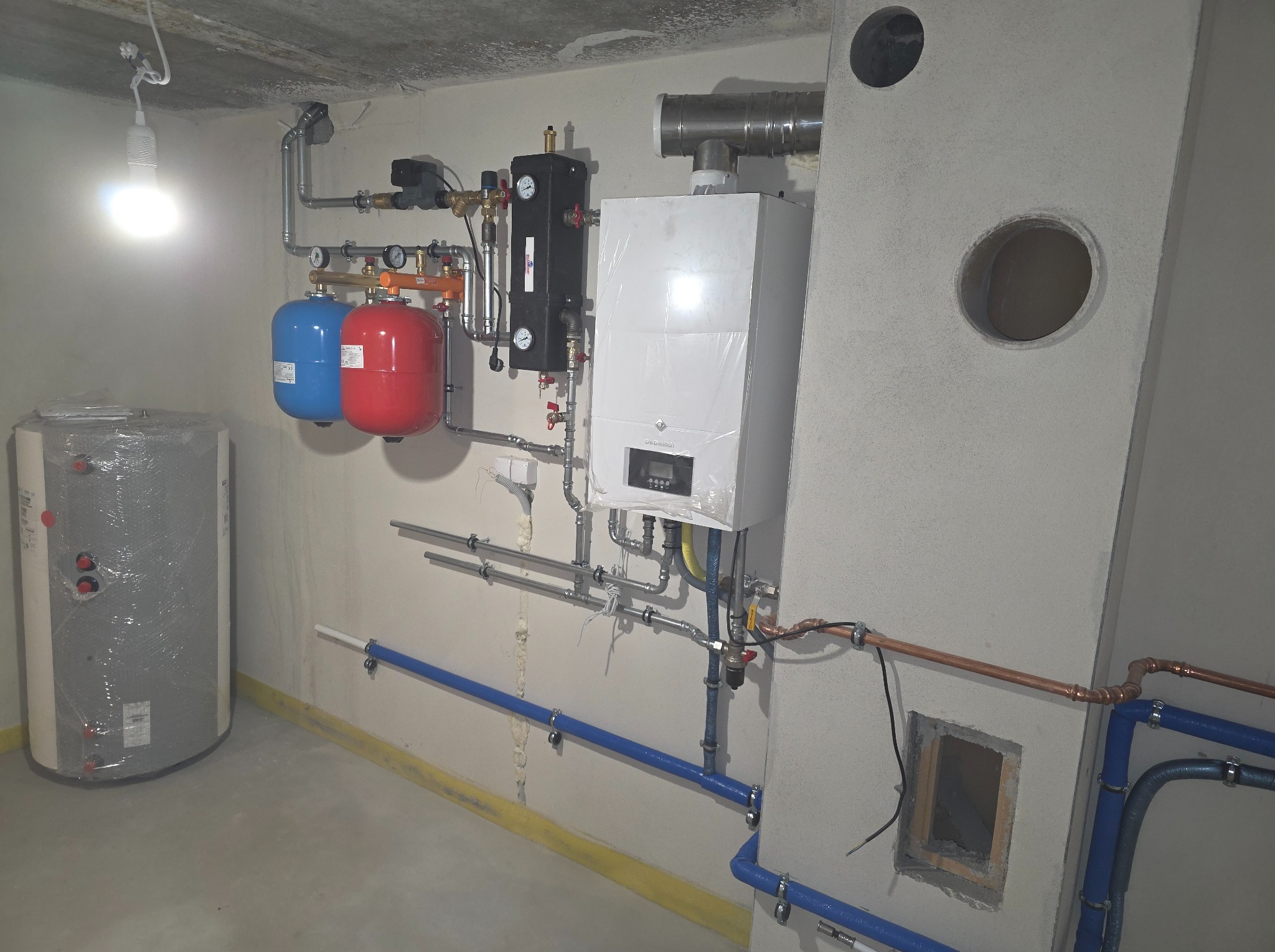 Złożona instalacja grzewcza z piecem, zbiornikiem i rurami w surowym pomieszczeniu. Widoczne elementy instalacji w kolorach niebieskim, czerwonym i miedzianym.