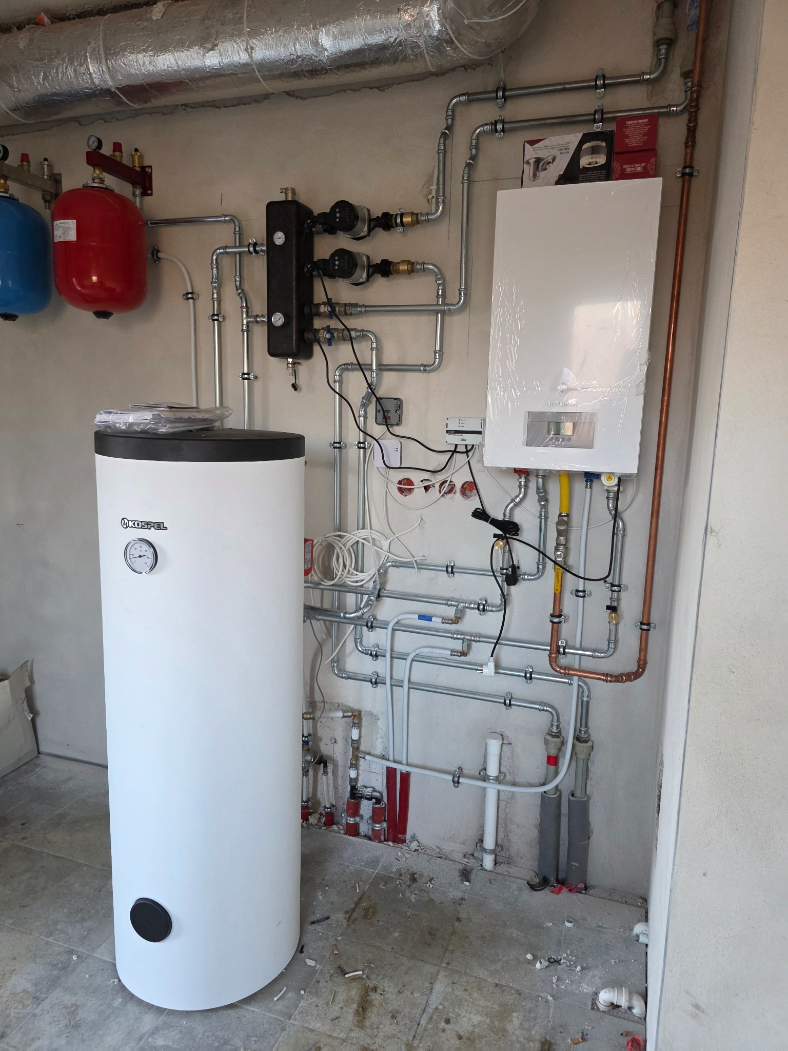 Biały zasobnik ciepłej wody Kospel z manometrem, obok piec CO w folii i instalacja grzewcza z rur miedzianych i stalowych na szarej ścianie.