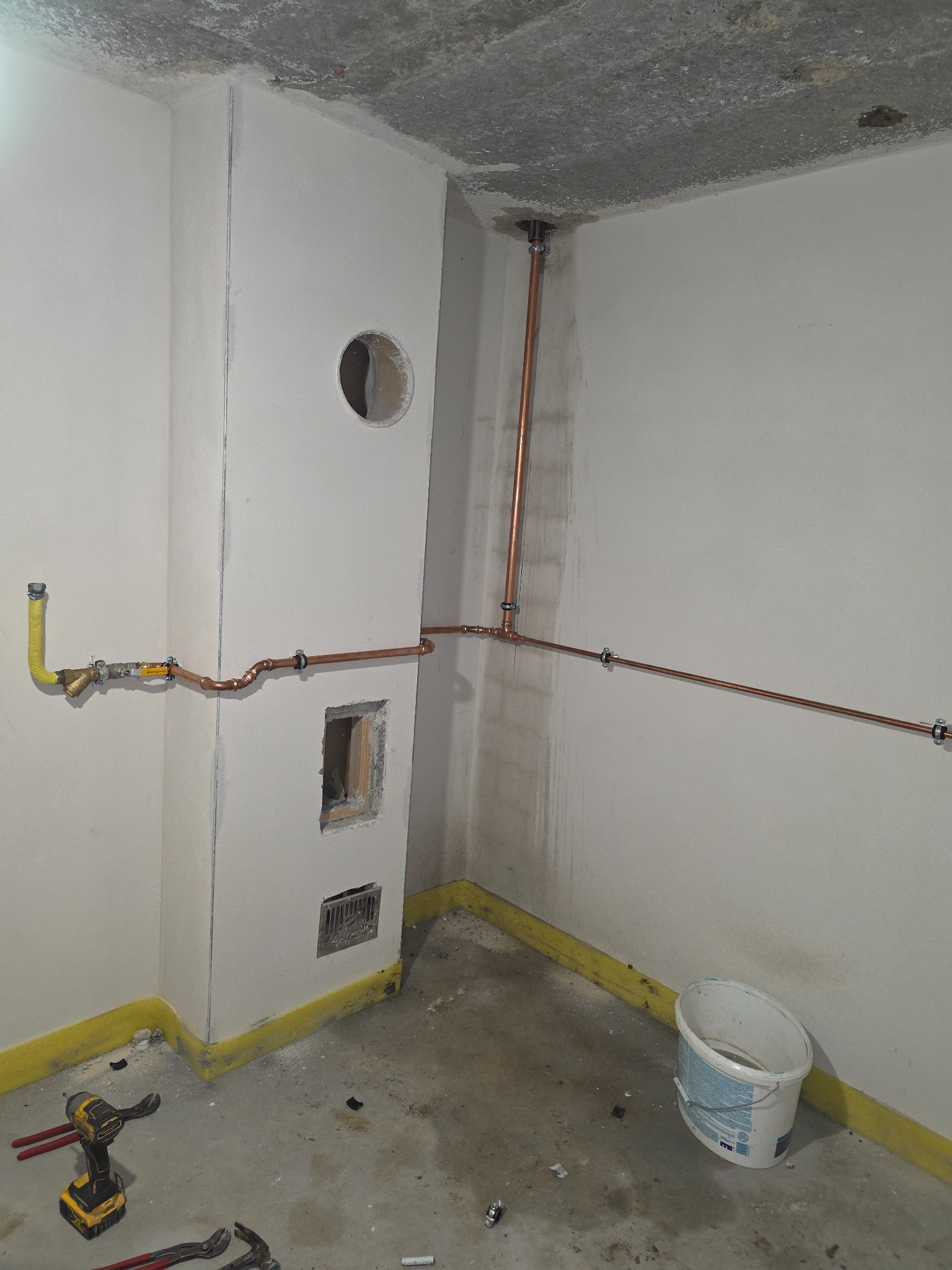 Miedziane rury instalacji gazowej zamontowane w surowym wnętrzu, z żółtym przewodem gazowym i narzędziami na podłodze. Widoczne otwory w ścianie i surowy betonowy sufit.