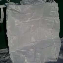 Biały, pusty worek typu Big Bag z uchwytami, leżący na śniegu przed zielonym metalowym tłem.