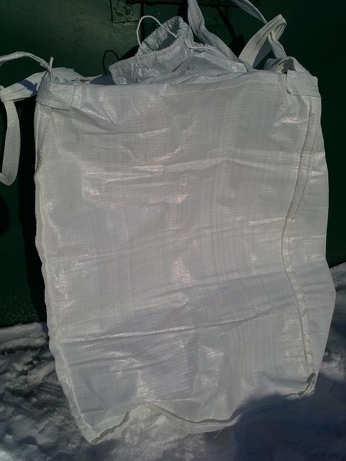 Biały, pusty worek typu Big Bag z uchwytami, leżący na śniegu przed zielonym metalowym tłem.