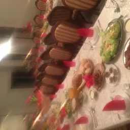 Długi stół nakryty białym obrusem z wzorem, zastawiony naczyniami, sztućcami i potrawami, takimi jak sałatki, pieczywo, wędliny i smażone mięso, z czerwonymi serwetkami na talerzach i bukietami...