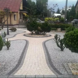 Ogród z krętą ścieżką z jasnej kostki brukowej obramowaną szarym kolorem, wysypaną drobnymi kamykami, z ozdobnymi krzewami i drzewkami, w tle dom jednorodzinny.