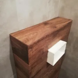 Obudowa stelaża WC z forniru drewnopodobnego z widocznym otworem rewizyjnym, na tle płytek w odcieniach szarości.