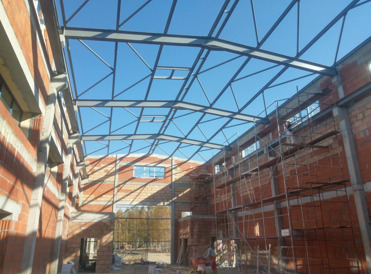 Wnętrze budowy hali: konstrukcja stalowa dachu, ściany z czerwonej cegły, rusztowania i pracownik budowlany z taczką na dole.