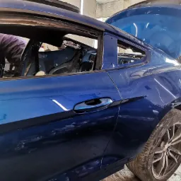 Niebieski samoch&oacute;d sportowy w trakcie naprawy blacharskiej, widoczne otwarte drzwi i bagażnik, pracownik wewnątrz pojazdu, brak szyby w drzwiach, koło z zabrudzeniami.